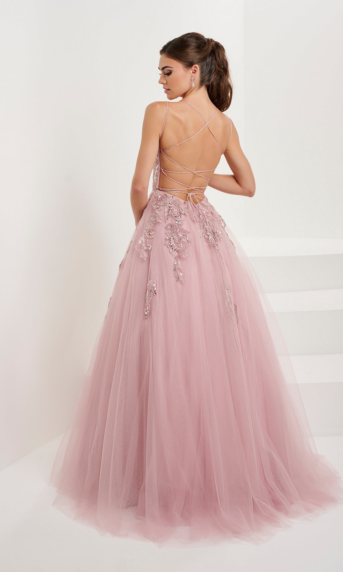 A woman wears the Tiffany Backless Tulle Ball Gown Prom Dress 16072 in light pink, featuring lace-up straps, sequin beaded embroidery, and a backless design, while standing in a minimal white room.